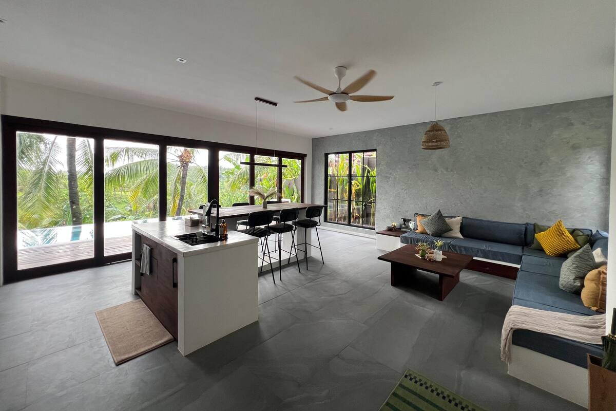 A spacious living area with a kitchen, large windows, and a ceiling fan.