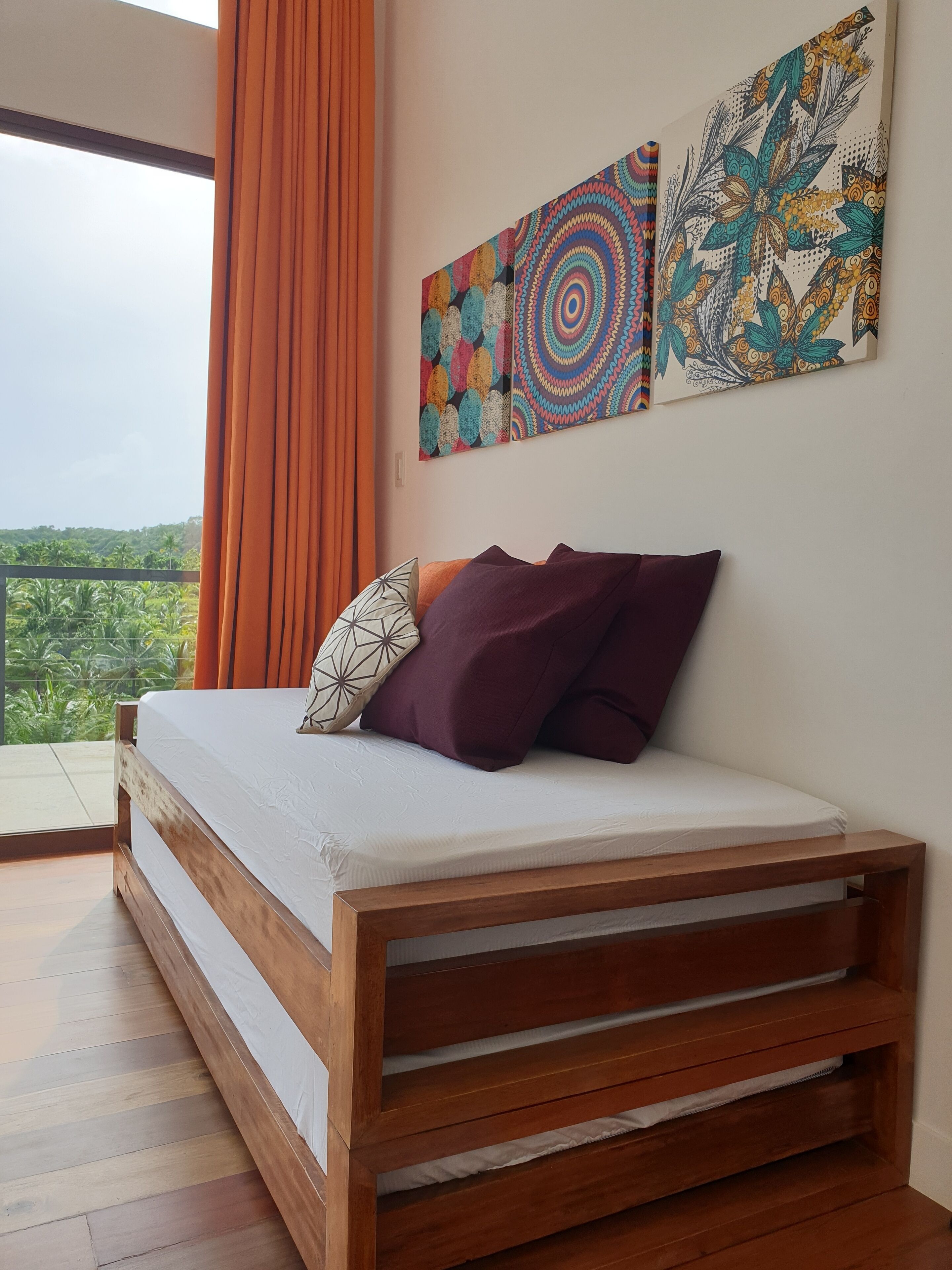 Quarto com cama de madeira, cortinas laranja e duas obras de arte coloridas nas paredes.