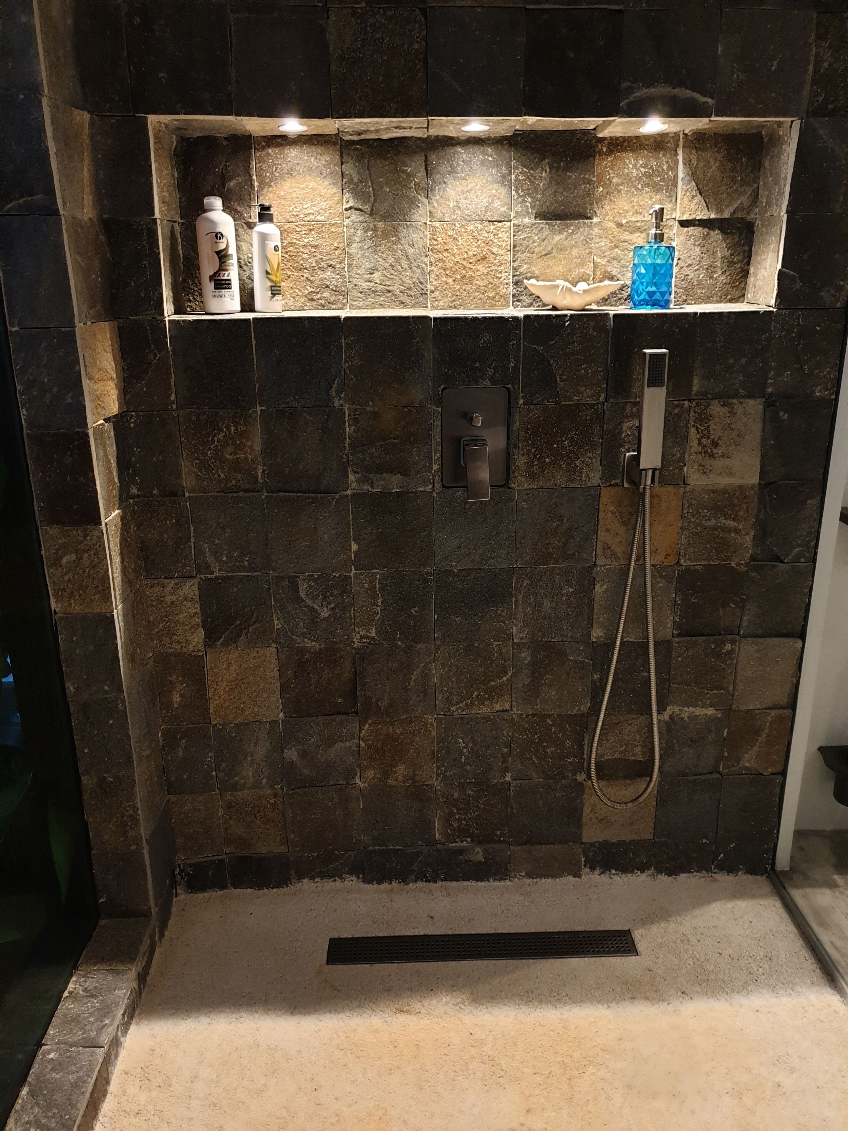 A bathroom with a stone wall, a shower, and a shelf with toiletries.