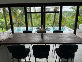 A modern dining area with a large table and three chairs, overlooking a pool and lush greenery.
