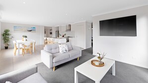 A modern living room with a grey sofa, a dining table, and a kitchen in the background. - Island Luxe Retreat Cowes. Modern Holiday House, Powerful outdoor spa/jaquzzi. (Cowes)