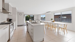 A modern kitchen with white cabinetry, a central island, and a dining area with white chairs. - Island Luxe Retreat Cowes. Modern Holiday House, Powerful outdoor spa/jaquzzi. (Cowes)