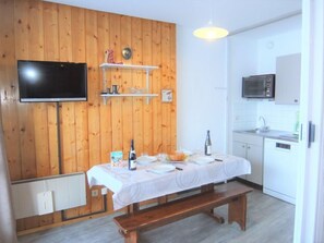 Una habitación acogedora con pared de madera, televisor de pantalla plana, mesa de comedor dispuesta con platos y una botella, y una zona de cocina con microondas y fregadero.