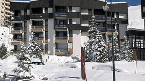 A modern multi-story building with balconies, surrounded by snow-covered trees and a clear sky.