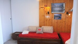 A small, cozy room with wooden walls, a bed with red bedding, a nightstand, and a wall-mounted shelf with a picture.