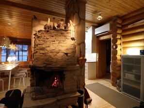 A stone fireplace with a fire, wooden ceiling, and a dining area with chairs and a table.