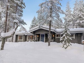 A log cabin with a snow-covered roof and multiple windows, surrounded by snow-laden trees.
