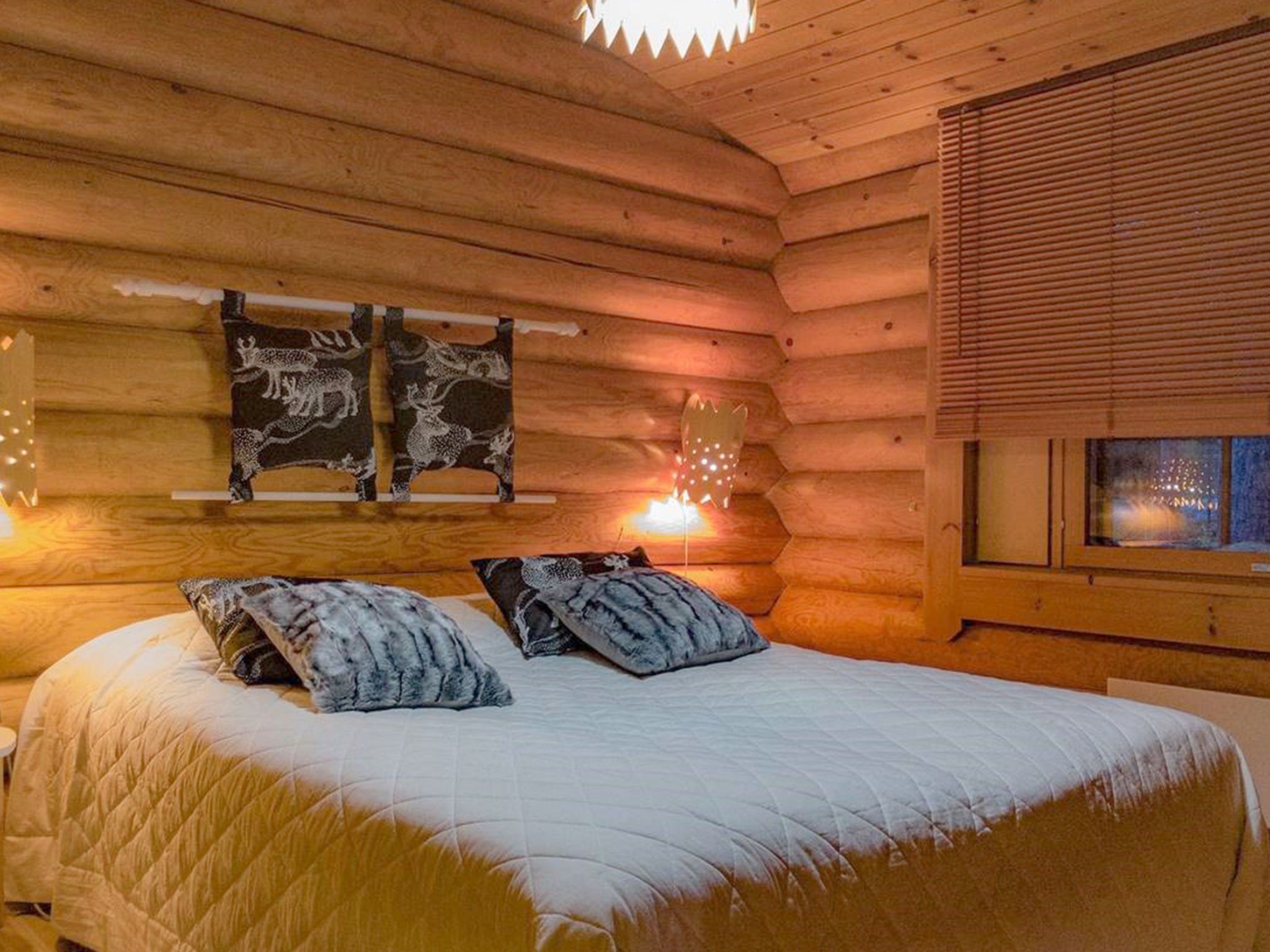 A log cabin bedroom with a bed, two pillows, a wall-mounted tapestry, and a window with blinds.