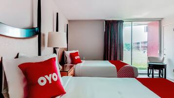 Habitación de hotel con dos camas, almohada roja con la marca OYO, mesita de noche con lámpara, una silla y vistas al exterior a través de una puerta corrediza.