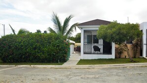 A residential house with a white exterior, a brown roof, and a small front yard with a palm tree and a bush with red flowers.