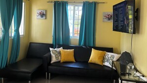 A living room with a black sofa, teal curtains, a ceiling fan, a TV, and a glass table with a remote.