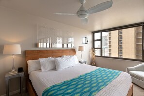 Une chambre d’hôtel avec un grand lit, un ventilateur de plafond, une fenêtre donnant sur des immeubles et deux lampes de chevet.