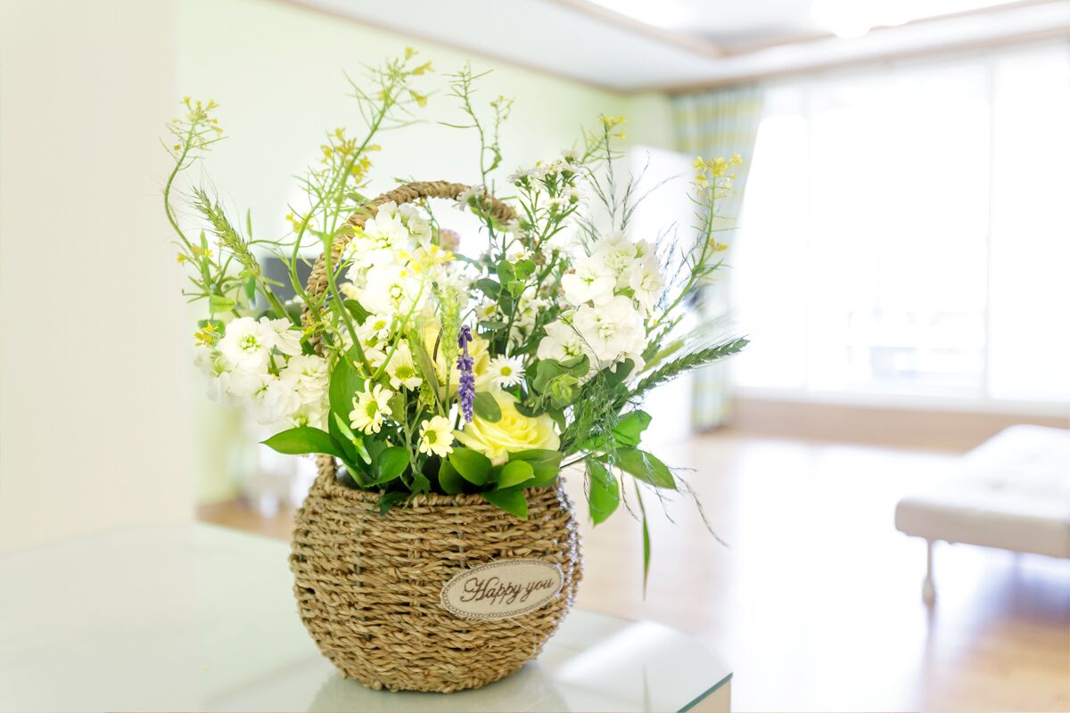 Una cesta de mimbre con la etiqueta 'Happy you', llena de flores blancas y amarillas, colocada sobre una mesa en una habitación luminosa con un ventanal grande.