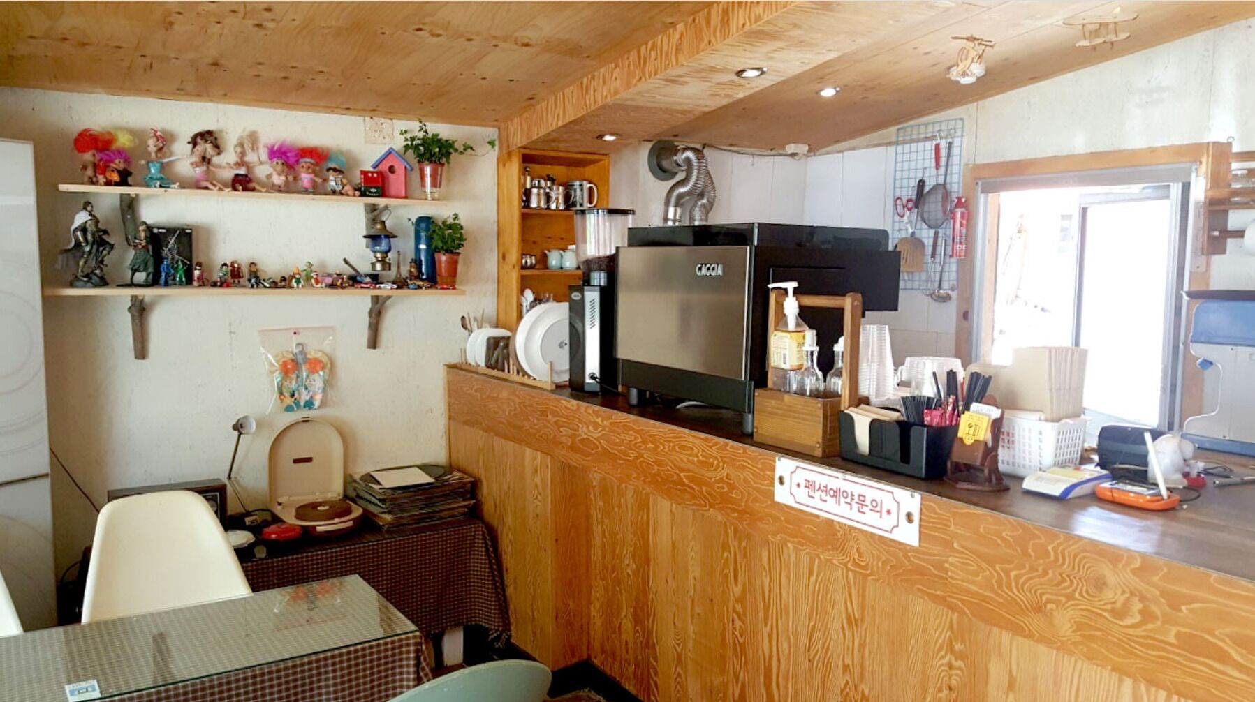 A small, cozy room with wooden walls and a wooden counter. There is a television on the counter, a small table with a chair, and shelves with various items.