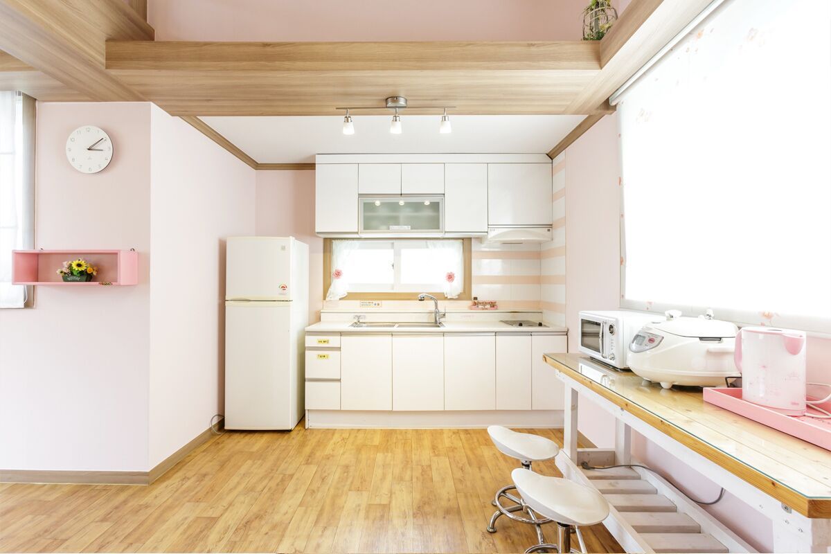 Cocina compacta con armarios blancos, un refrigerador, un microondas y un lavamanos. Hay un estante rosa pequeño en la pared y un taburete en la encimera.