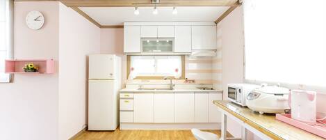 Cocina compacta con armarios blancos, un refrigerador, un microondas y un lavamanos. Hay un estante rosa pequeño en la pared y un taburete en la encimera.