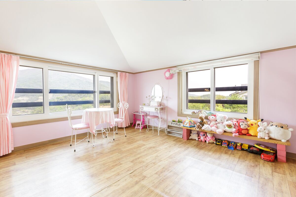 Una stanza dalle pareti rosa, pavimento in legno, una scrivania con una sedia, una finestra con vista sulle colline e uno scaffale con peluche.