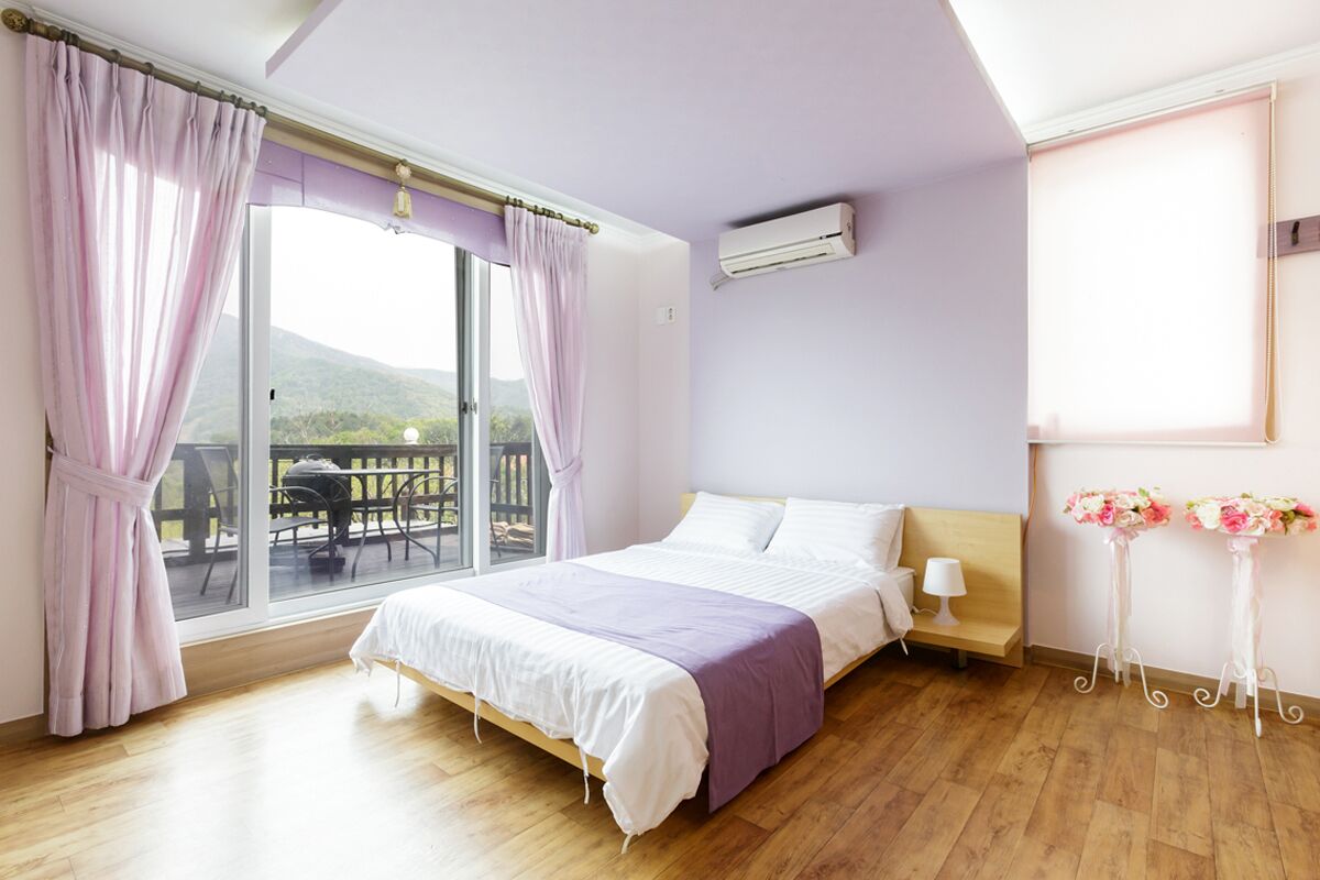 Un dormitorio con cama, ventana con cortinas, balcón, unidad de aire acondicionado y una pequeña mesa con lámpara.