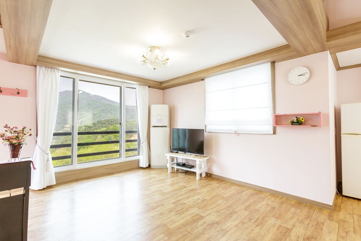 A spacious room with a large window, a flat-screen TV on a stand, a white refrigerator, and a wooden ceiling.