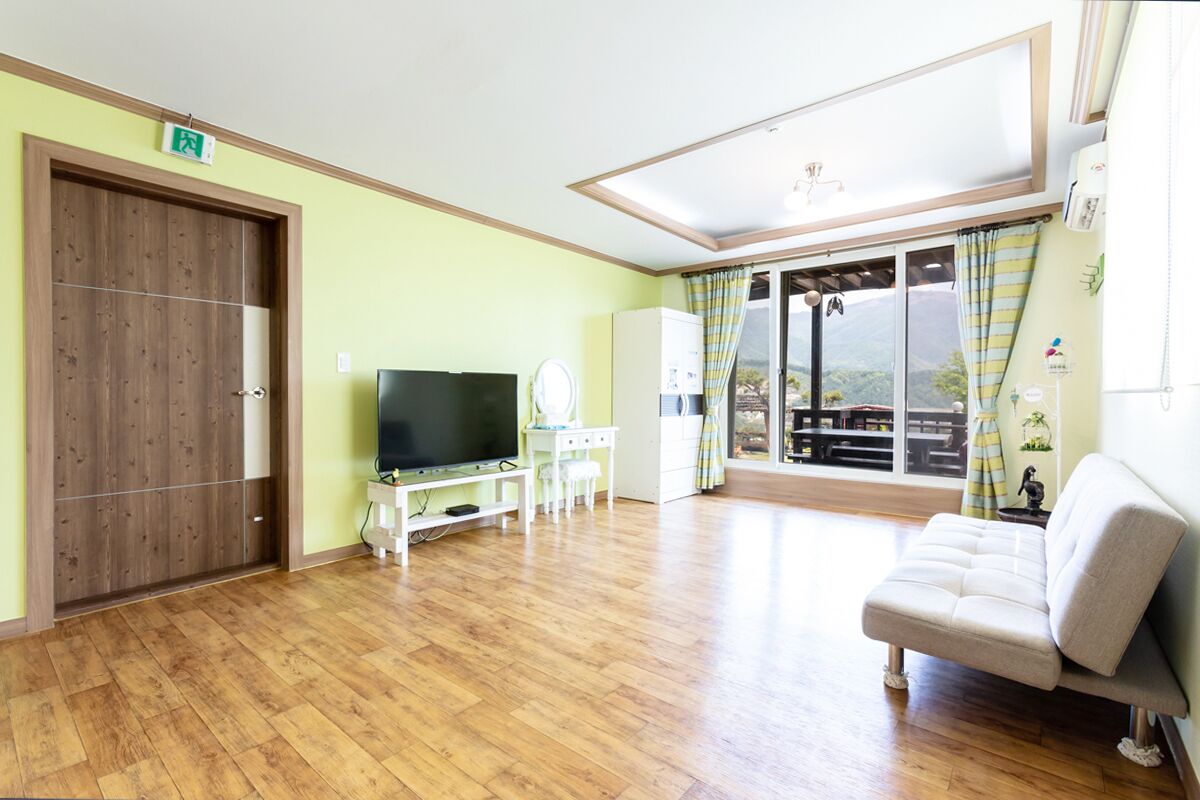 A room with a wooden floor, a television, a sofa, a desk with a chair, and a window with curtains.