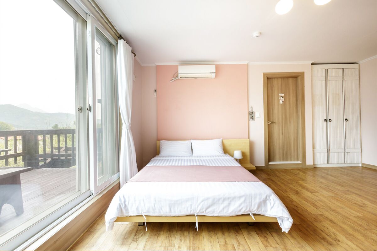 A bedroom with a large window, a bed, a wardrobe, and an air conditioning unit.