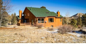A two-story log cabin with a green roof, a front porch, and a view of a mountain in the background.