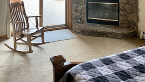 A cozy room with a stone fireplace, wooden ceiling, and a rocking chair near a sliding glass door leading to an outdoor deck.