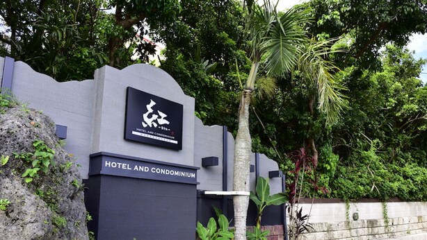 A sign for a hotel and condominium with a logo, surrounded by lush greenery and a stone wall. - Hotel Bin (Okinawa)