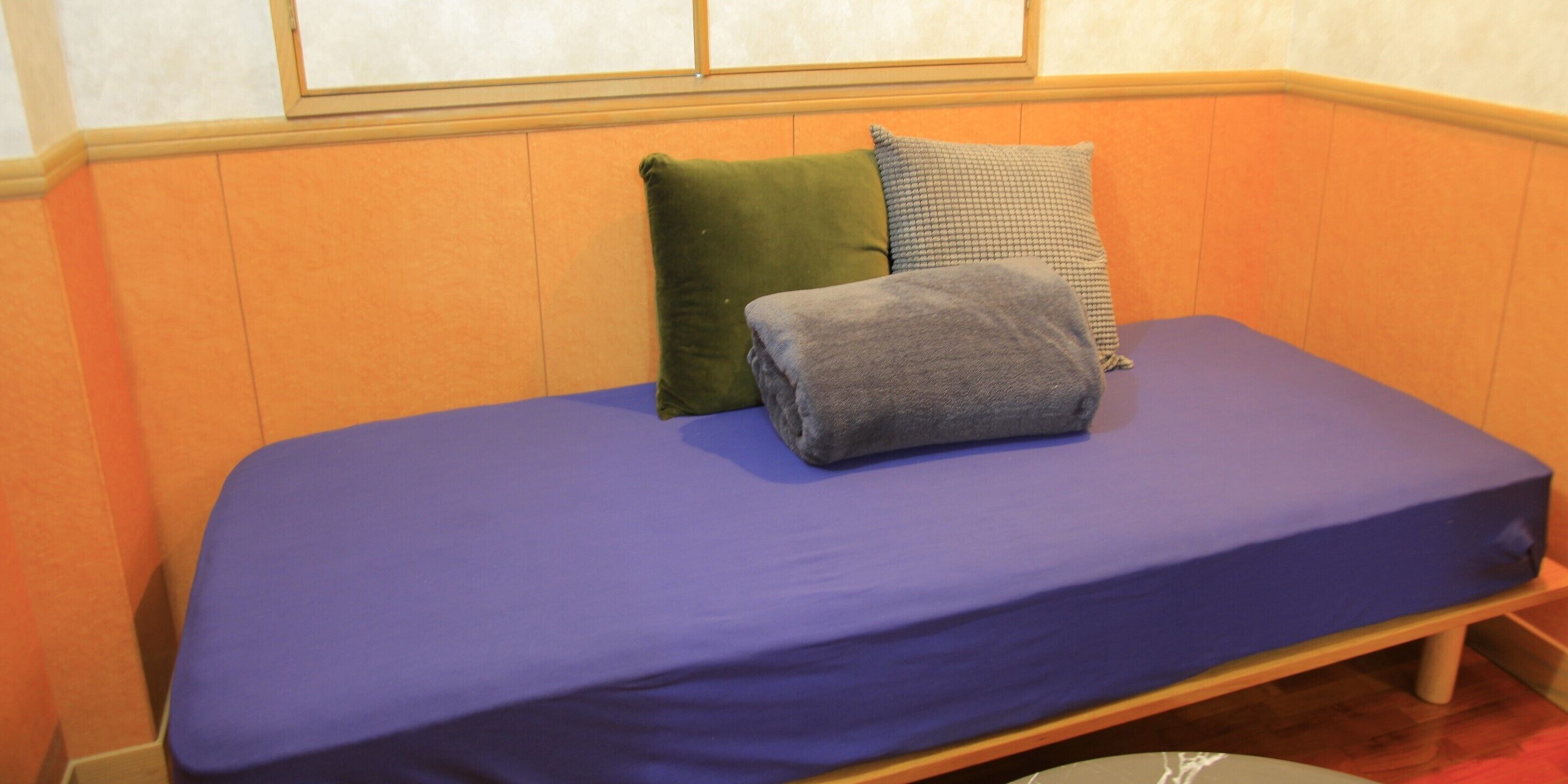 Une petite chambre avec un lit simple, un matelas violet, deux oreillers et un banc en bois.