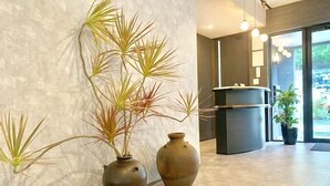 A reception area with a modern design, featuring a reception desk, a large window with a view of greenery, and decorative plants including a tall palm and two vases. - Hotel Bin (Okinawa)
