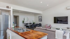 Cocina moderna con encimera de madera, lavaplatos integrado y refrigerador de acero inoxidable. Hay una zona de comedor con mesa y sillas, y una sala de estar con sofá y televisor montado en la pared.