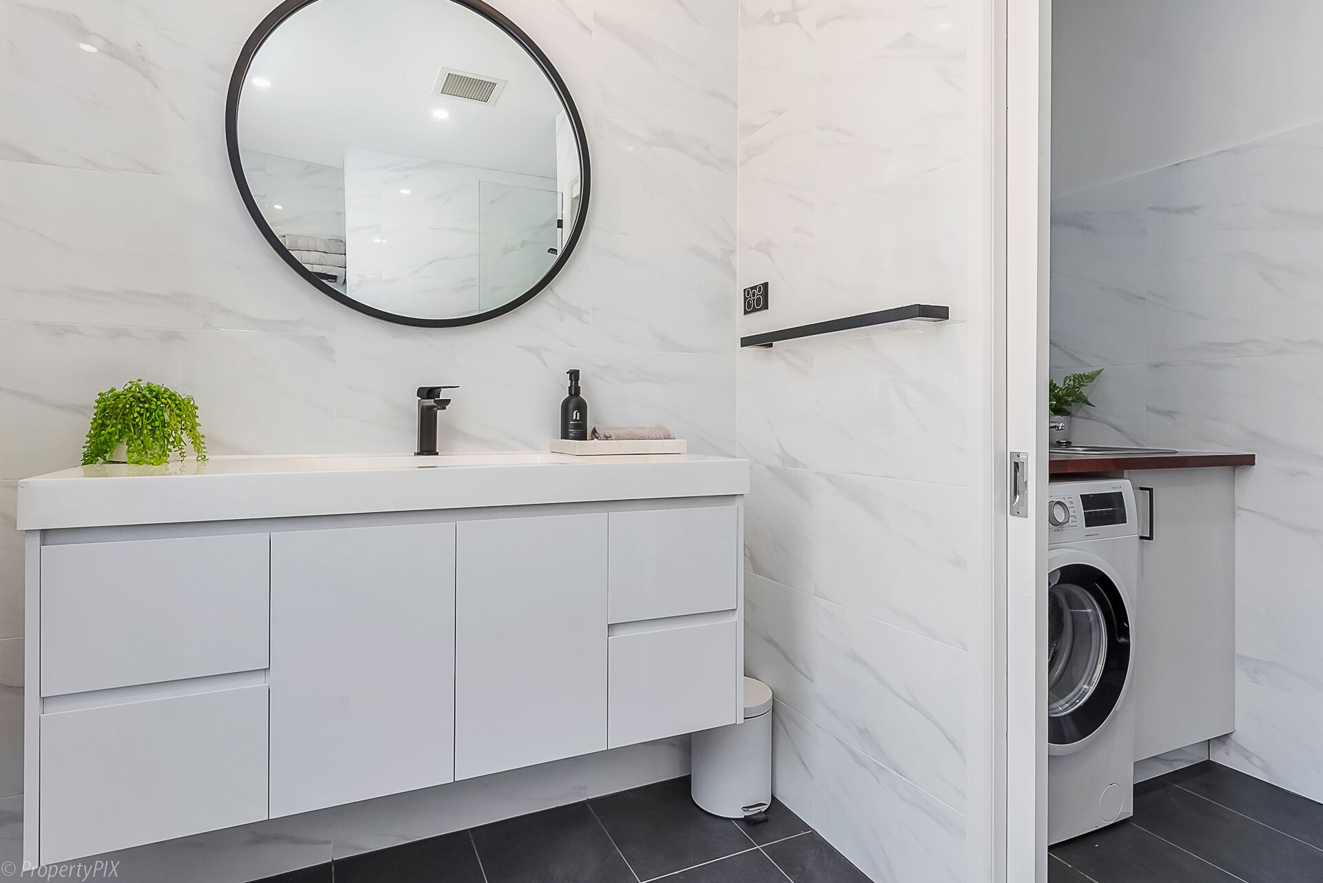 A modern bathroom with a white marble vanity, a round mirror, a black faucet, and a built-in washing machine.