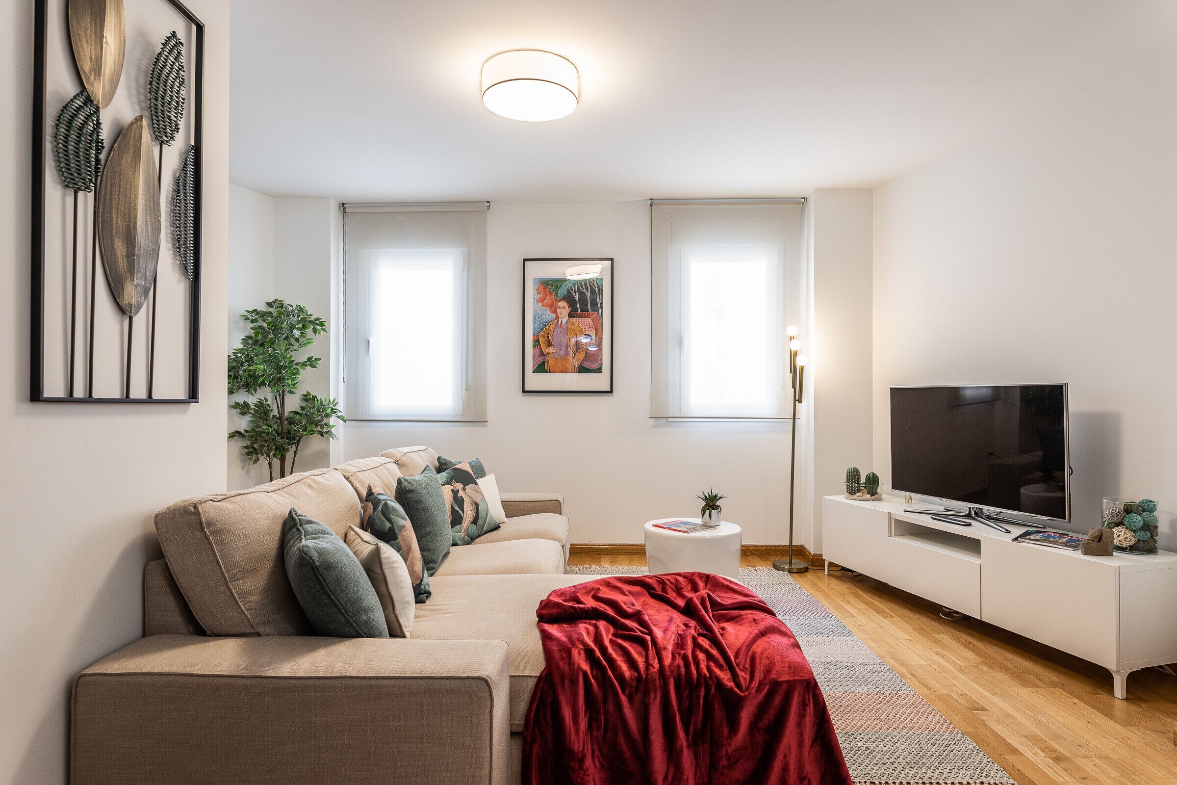 Un salon moderne avec un canapé beige, un plaid rouge, un téléviseur à écran plat, un meuble de rangement blanc et un tableau encadré accroché au mur.