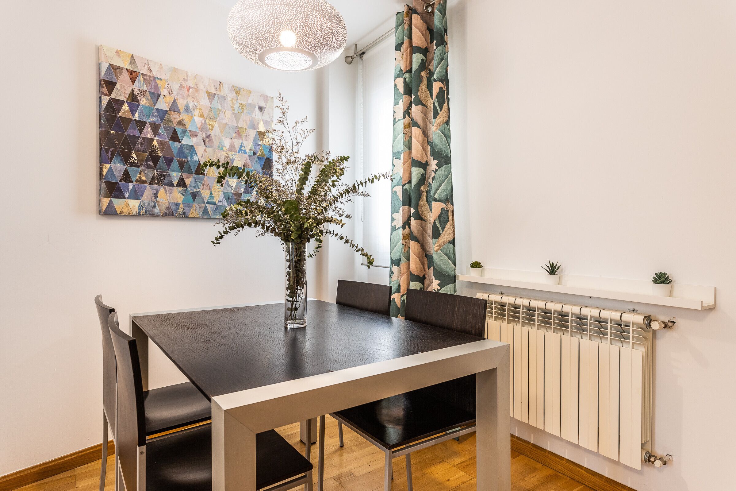 A dining area with a table, chairs, a vase with plants, a geometric artwork, a window with curtains, and a radiator.