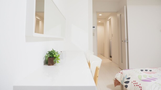 Una stanza minimalista con un letto bianco, un tavolino con una pianta e uno specchio al muro.