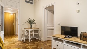 Ein Zimmer mit Holzdielen-Fußboden, einem Fernseher auf einem Holztisch, einem kleinen Tisch mit Stühlen, einer Vase mit Pflanzen und einem Kleiderschrank.