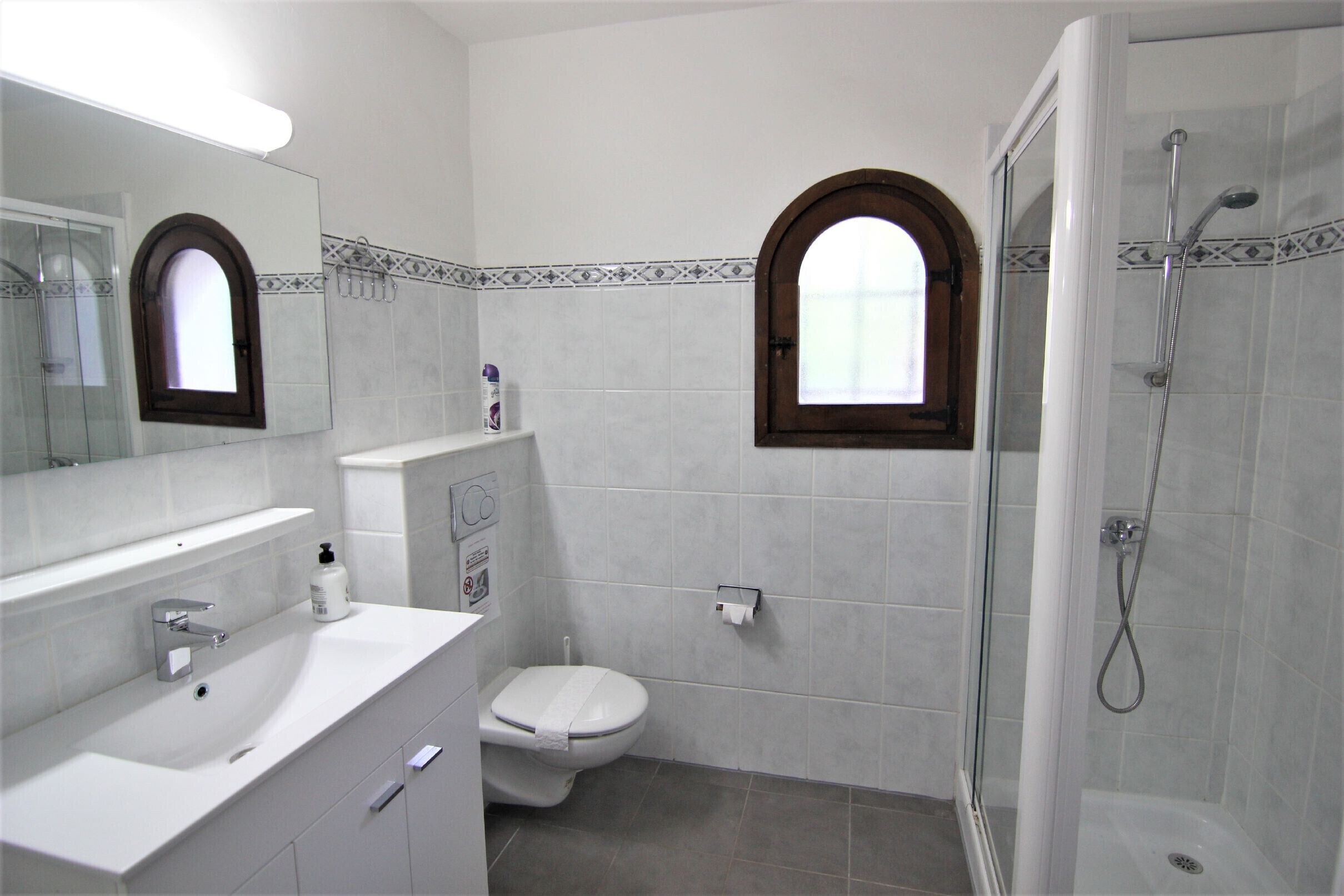 Une salle de bain avec un lavabo blanc, des toilettes, une douche et deux fenêtres en arc de cercle.