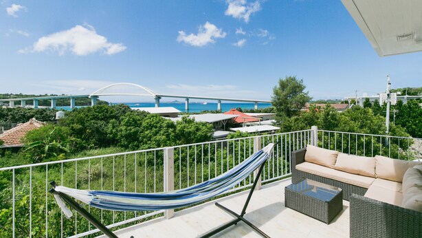 A balcony with a hammock, wicker sofa, and a view of a bridge and water. - Sea Sunset Villa (Motobu)
