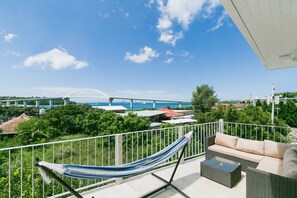 A balcony with a hammock, wicker sofa, and a view of a bridge and water. - Sea Sunset Villa (Motobu)