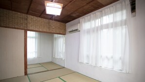 Ein Zimmer mit Tatami-Boden, weißen Wänden, einem Deckenventilator und einem Fenster mit durchsichtigen Vorhängen.