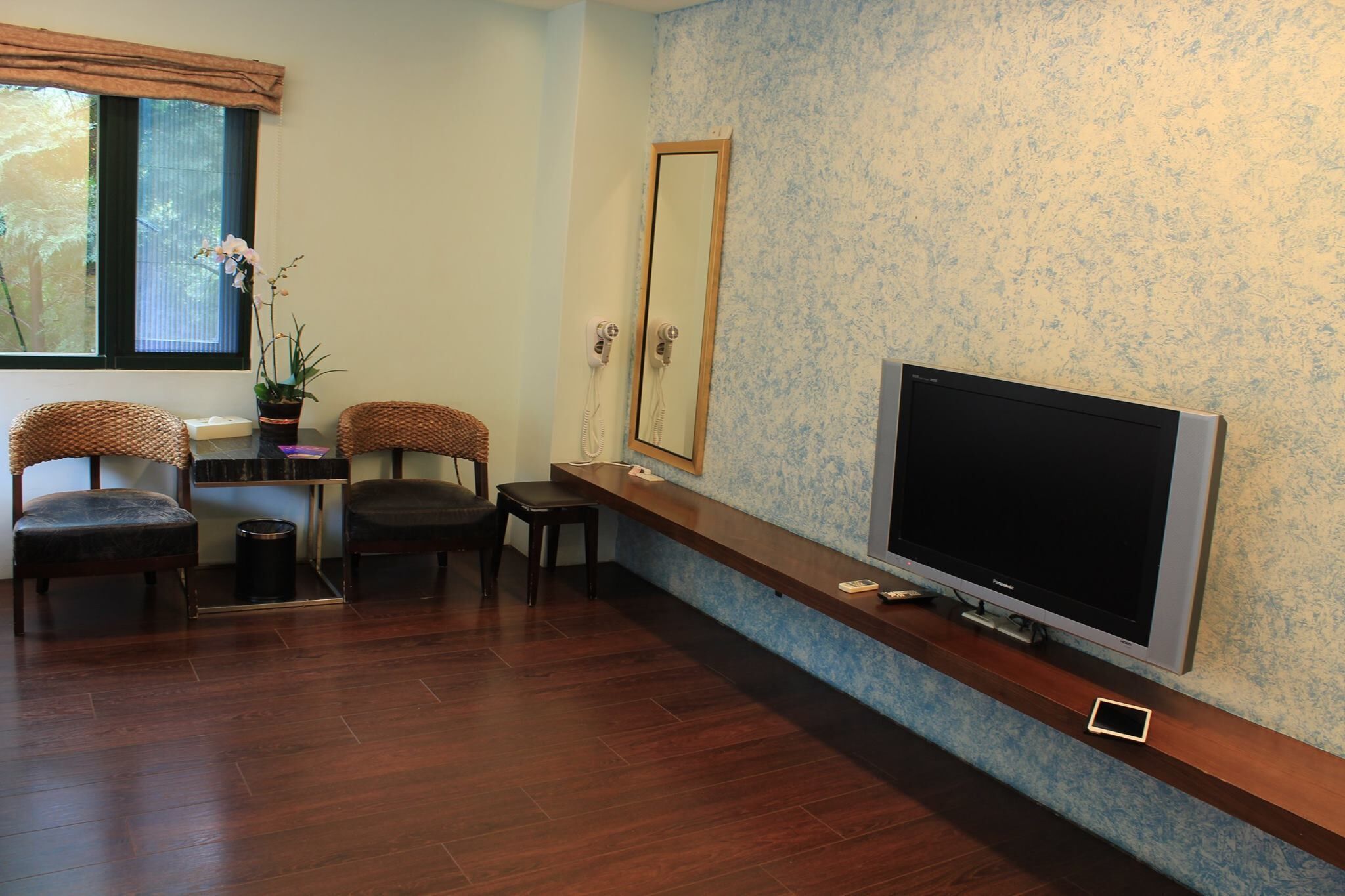A room with a flat-screen TV, two wicker chairs, a small table, a potted orchid, and a mirror on the wall.