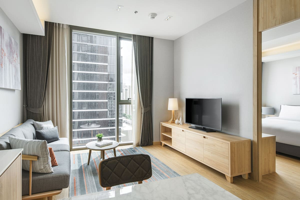 Ein modernes Hotelzimmer mit einer Couch, einem Fernseher, einem Holzschrank, einem kleinen Tisch mit Lampe und einem großen Fenster mit Vorhängen.