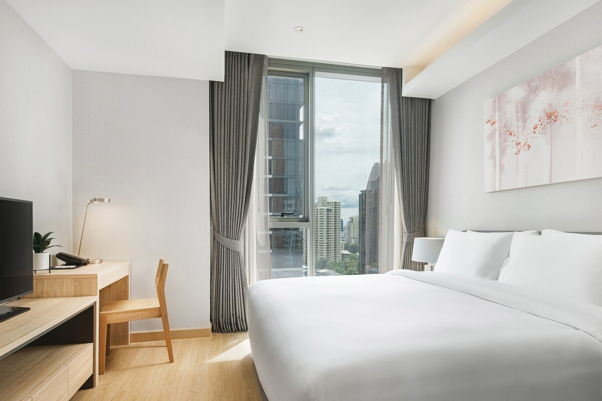 Une chambre d’hôtel avec un lit, un bureau, une chaise, une télévision et une grande fenêtre avec des rideaux.