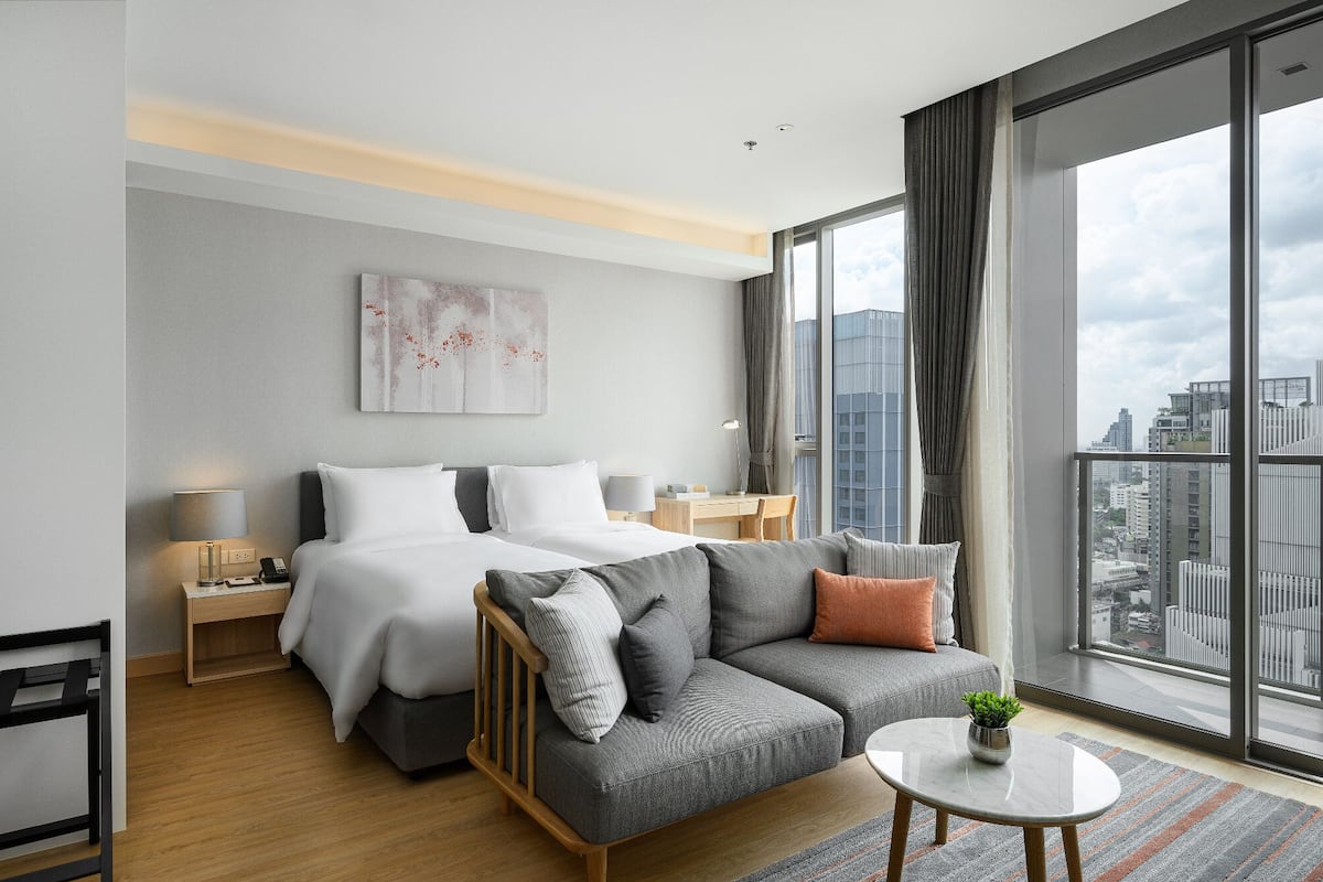 Ein modernes Hotelzimmer mit einem großen Bett, einer Sitzecke, einem kleinen Tisch und Blick auf die Stadt durch große Fenster.