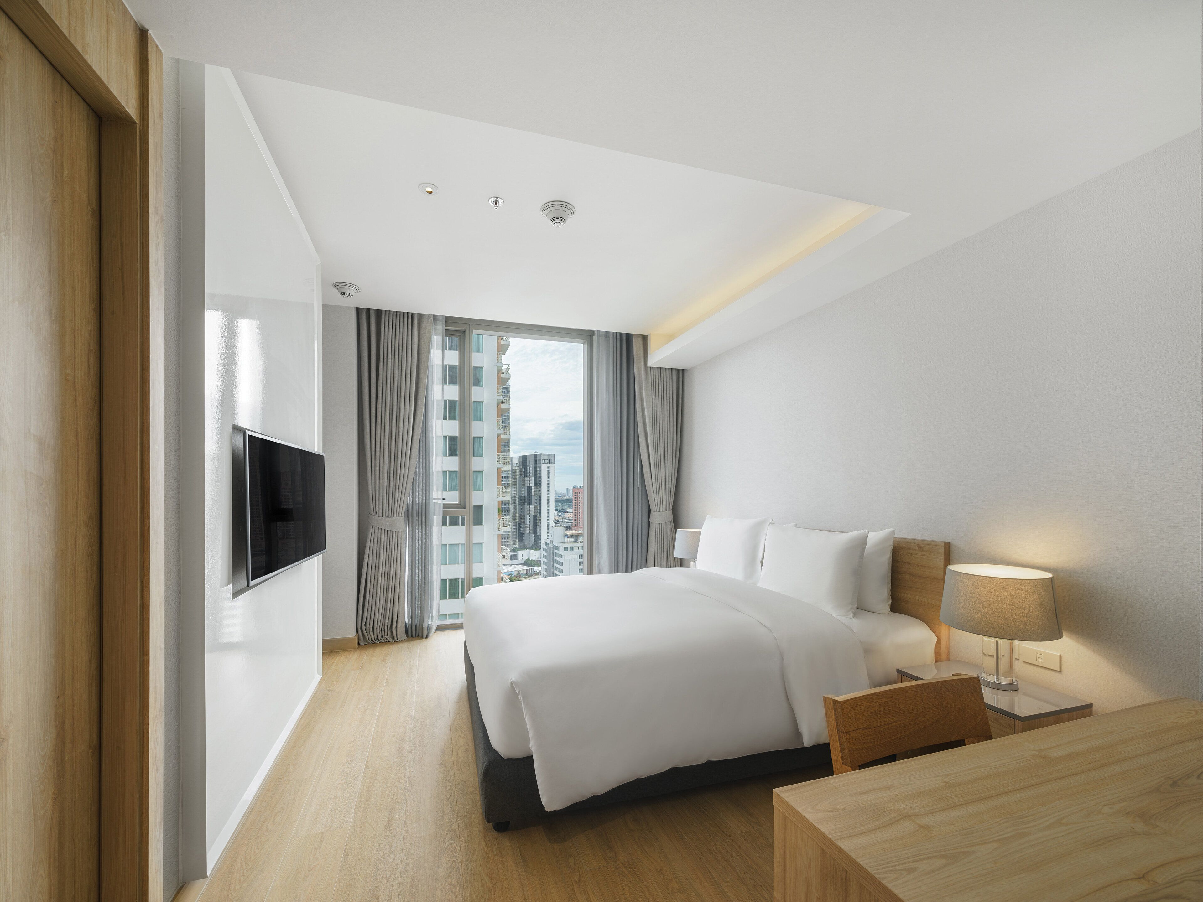 A hotel room with a bed, a TV mounted on the wall, a desk with a lamp, and a view of the city through the window.