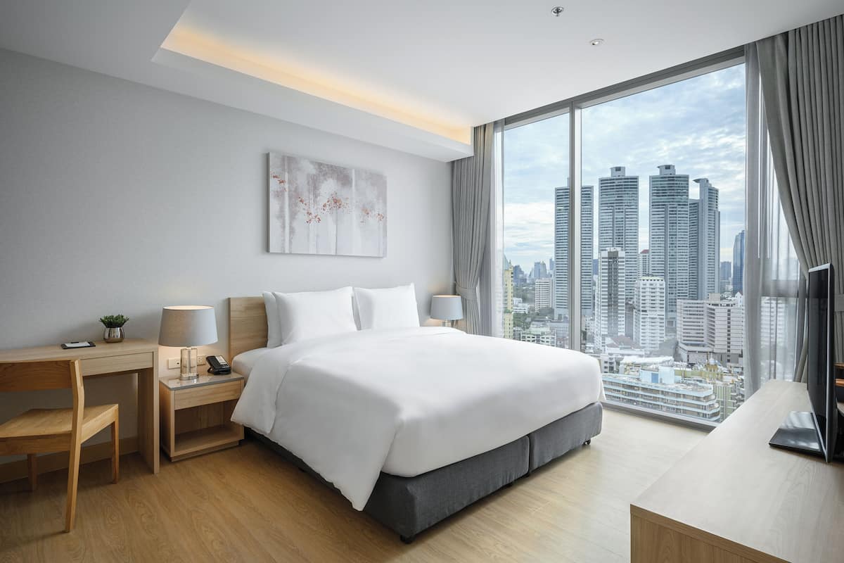Ein modernes Hotelzimmer mit einem großen Bett, einem Schreibtisch, einem Sessel, einem Nachttisch mit Lampe und Blick auf die Stadt.