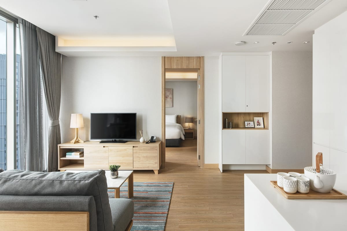 Ein modernes Hotelzimmer mit Fernseher, einer Couch, einem Essbereich mit Tisch und Stühlen sowie Blick auf die Stadt durch große Fenster.
