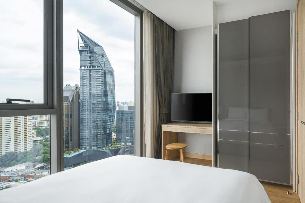 A modern hotel room with a large window, a bed, a desk with a chair, and a wardrobe.
