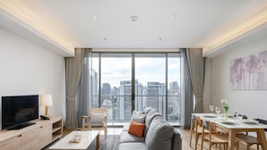 Een moderne woonkamer met een grote bank, een televisie, een eethoek met een tafel gedekt voor twee personen, en uitzicht op de stad door grote ramen.
