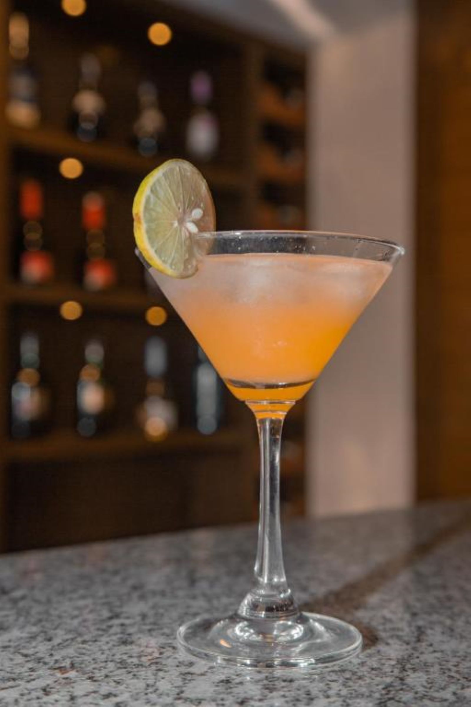 En cocktail i ett martiniglas, garnerad med en citronskiva, står på bardisken.
